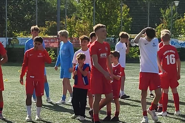 Mehrere Jugendliche in roten Fußballtrikots stehen auf einem Kunstrasenplatz. Einige sind in Gruppen versammelt.
