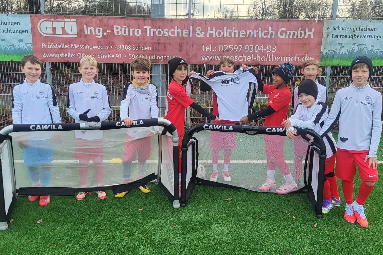 Gruppe von Kindern in Fußballtrikots steht an einem Kunstrasenplatz mit Minitoren.